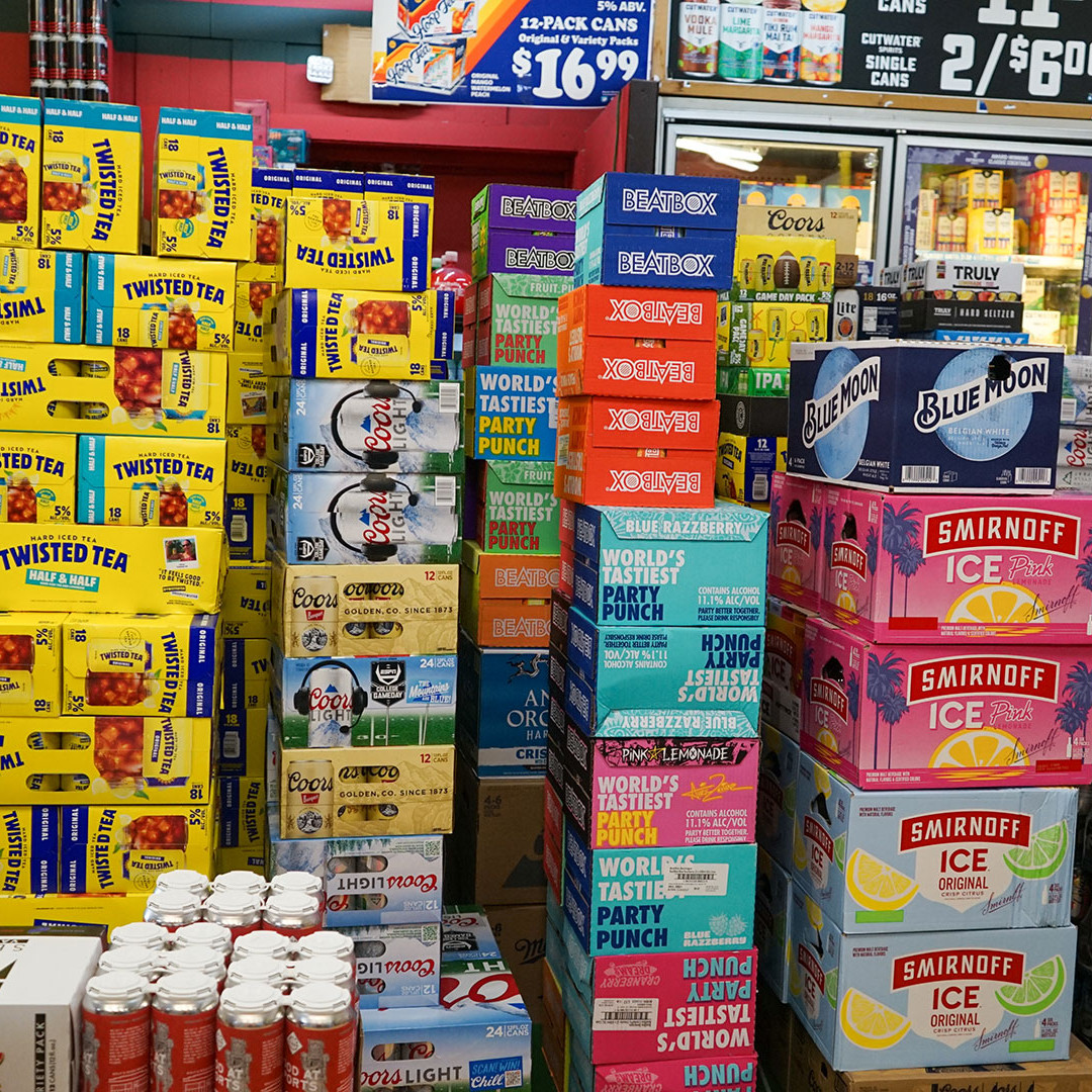 A display of various beverage boxes stacked high in a store, including brands like Twisted Tea, Coors Light, BeatBox, Blue Moon, and Smirnoff Ice, with a refrigerator section visible in the background.