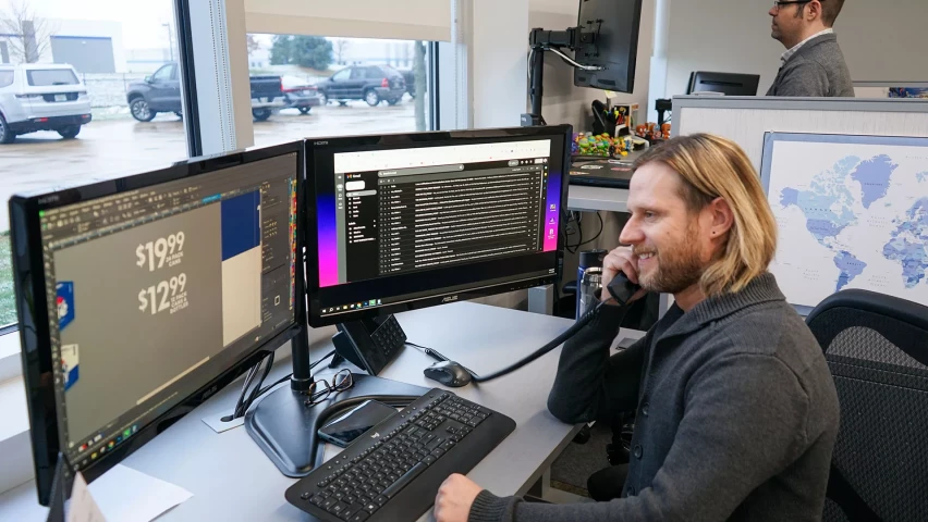 A smiling man with long blonde hair sits at a desk with dual monitors, talking on the phone in an office with large windows. One monitor shows email, while the other displays design software. Another worker is standing at a computer in the background.