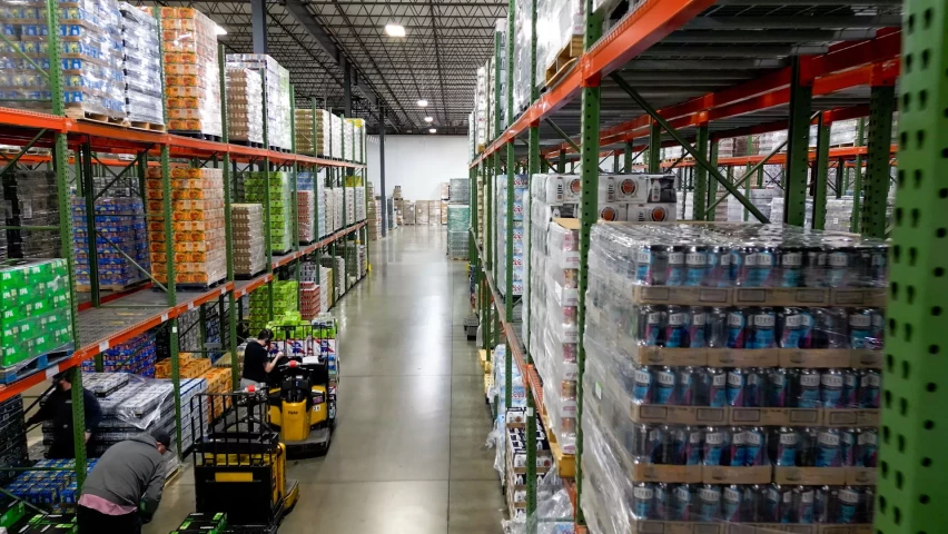 A large warehouse aisle filled with stacked pallets of beverages on both sides. Workers are seen in the aisle operating pallet jacks and organizing the inventory.