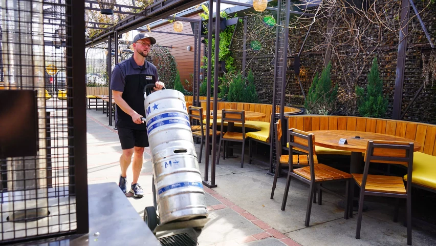 A worker in a dark uniform and cap transports a large silver keg on a hand truck through an outdoor seating area with wooden tables and chairs.