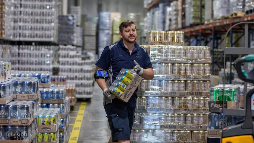 A warehouse worker in a blue uniform and gloves carries a case of canned drinks while navigating through an aisle filled with stacked pallets of beverages in a warehouse.