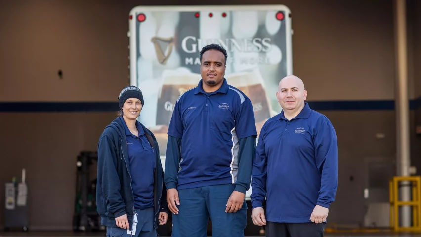 altThree warehouse workers in blue uniforms stand in front of a Guinness delivery truck, posing for a group photo.