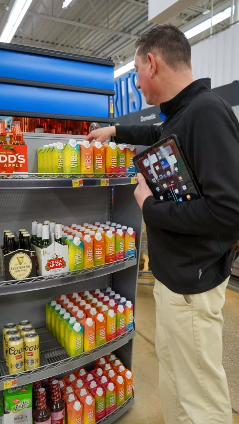 A store employee in a black jacket holds a tablet while arranging colorful Vibe drink cartons on a shelf, with other beverages like Guinness and Stella Artois visible on the lower shelves.