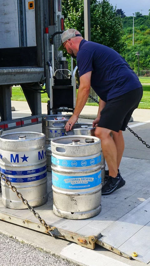 A worker in a blue shirt and shorts loading or unloading silver kegs from the back of a truck at a delivery site, with green grass and a handicapped parking sign visible in the background.