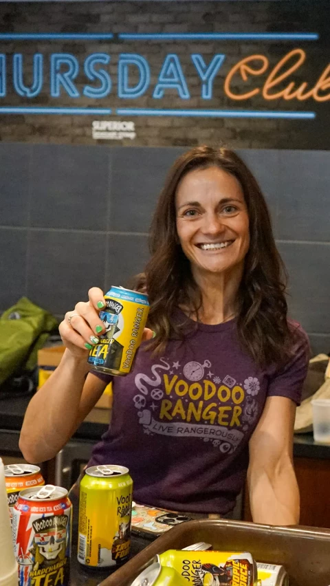 A smiling woman in a purple Voodoo Ranger shirt holds up a can of Voodoo Ranger beer, showing off her green-painted nails, with a background of a room and some belongings.