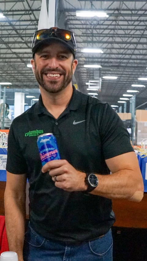 A smiling man in a black Columbus shirt and cap holds a can of Blueberry Shimmer beer while standing in a warehouse with industrial lighting and equipment in the background.