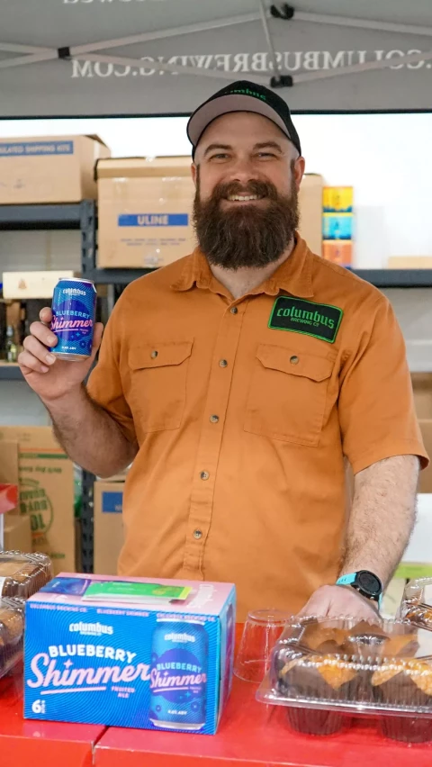 A bearded man in an orange shirt and cap, wearing a Columbus patch, smiles while holding a can of Blueberry Shimmer beer and standing behind a table with more beer cans and packaged muffins.