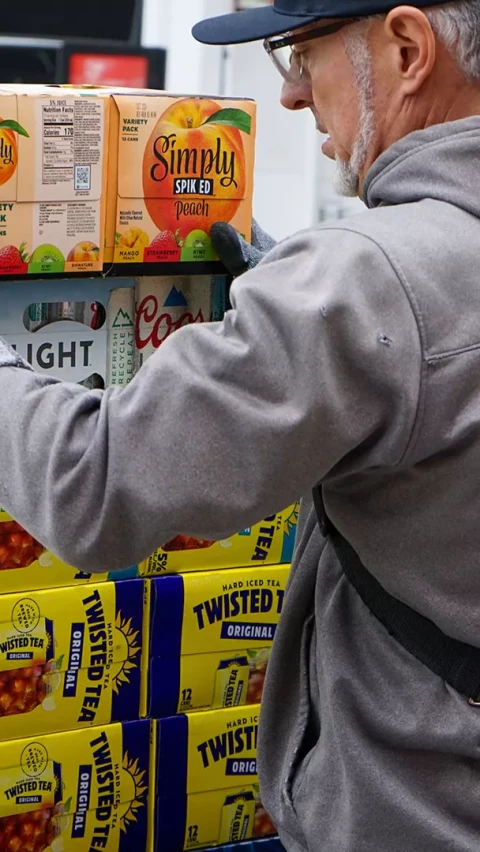 A worker in a grey jacket and cap stacks boxes of Simply Spiked Peach, Coors Light, and Twisted Tea in a store.