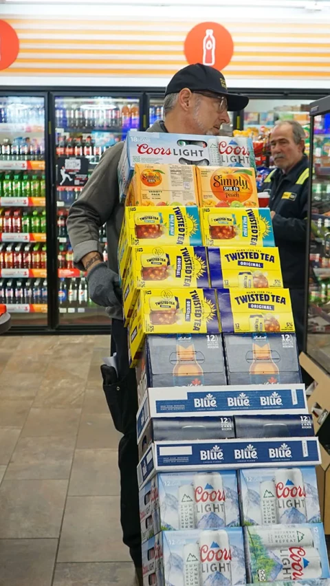 A worker in a convenience store moves a loaded hand truck stacked with boxes of Coors Light, Twisted Tea, Simply Spiked, and Labatt Blue, while another worker is visible in the background near the refrigerated beverage section.