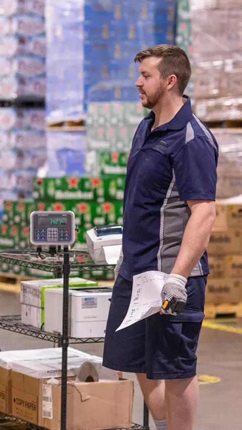 A worker in a blue uniform and gloves holds a paper and scanning device while standing in a warehouse aisle, with stacks of Heineken and other beverage boxes visible in the background.