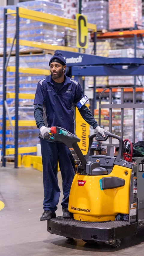 A warehouse worker in a blue uniform operates a yellow Jungheinrich pallet jack inside a warehouse, with shelves of goods in the background.