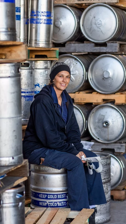 A woman in a dark hoodie and beanie sits on a keg in a warehouse filled with stacks of silver kegs, smiling at the camera.