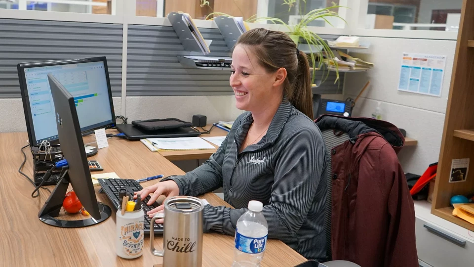 A smiling woman in a grey shirt sits at a desk typing on a keyboard in an office cubicle, with a computer monitor, a laptop, and various office supplies around her.